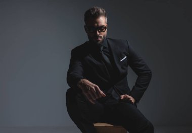 Siyah takım elbiseli ciddi dramatik bir adam otururken koyu gri arka planda güneş gözlüğü takıyor.