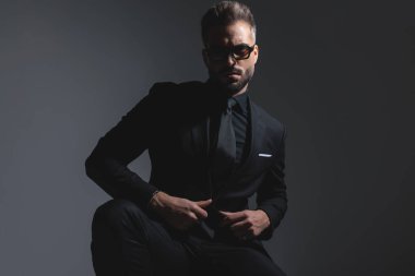 Siyah takım elbiseli cüretkar bir adam güneş gözlüğü takıyor ve koyu gri arka planda ileriye bakıyor.