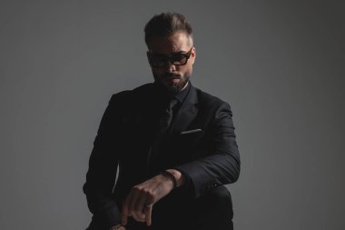 Siyah takım elbiseli koyu renkli bir adam güneş gözlüğü takıyor ve koyu gri arka plana bakıyor.