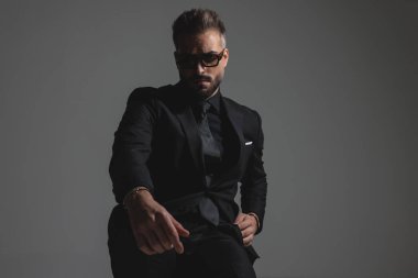 Koyu gri arka planda otururken siyah takım elbiseli ve güneş gözlüklü karamsar dramatik adam arkasına yaslanıyor.