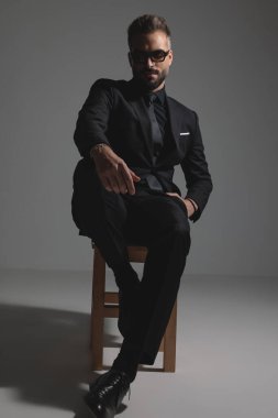 Siyah takım elbiseli ve güneş gözlüklü kendine güvenen şık adam oturuyor ve koyu gri arka plana yaslanıyor.