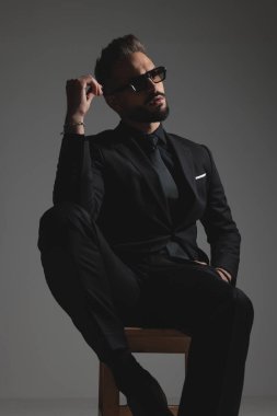 Koyu gri arka planda güneş gözlüklerini ayarlarken takım elbiseli düşünceli adam taburede oturuyor.