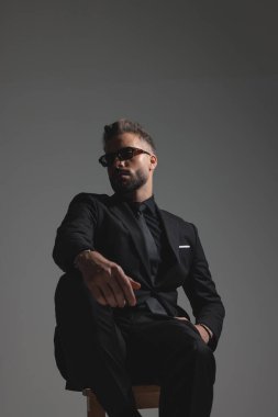 Siyah takım elbiseli ve güneş gözlüklü havalı adam arkasını yaslıyor ve koyu gri arka plana bakıyor.