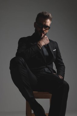 Meraklı cüretkar takım elbiseli adam taburede oturuyor ve koyu gri arka planda çeneye dokunuyor.
