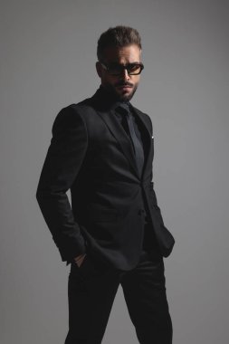 Takım elbiseli ve güneş gözlüklü ciddi bir moda adamı elleri ceplerinde koyu gri arka planda duruyor.