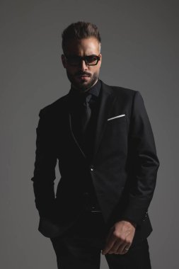 Siyah takım elbiseli esrarengiz esmer adam ve güneş gözlüğü koyu gri arka planda el ele poz veriyor.