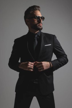 Siyah takım elbiseli, güneş gözlüklü şık bir adam ceketini ilikliyor ve koyu gri arka plana bakıyor.