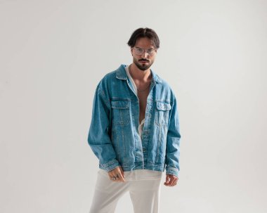 Kot ceketli ve beyaz pantolonlu şık bir adam açık gri arka planda poz veriyor ve ileriye bakıyor.