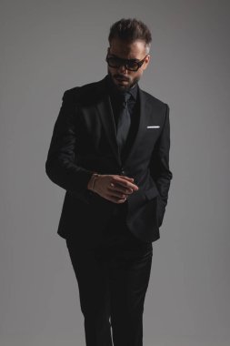 Modern takım elbiseli ve güneş gözlüklü adam koyu gri arka planda el ele yürüyor.