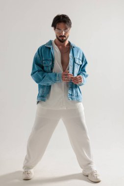 Kot ceketli şık bir adamın, parmaklarını bir arada tutarken ve açık gri arka planda ileriye bakarken tüm vücudunun fotoğrafı.