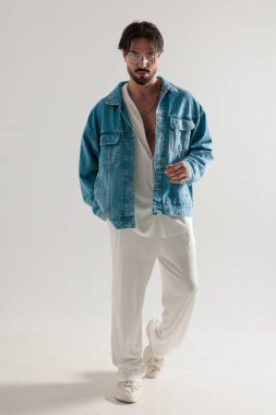 Kot ceketli ve beyaz giysili modacı ileri doğru yürüyor ve açık gri arka planda ileriye bakıyor.