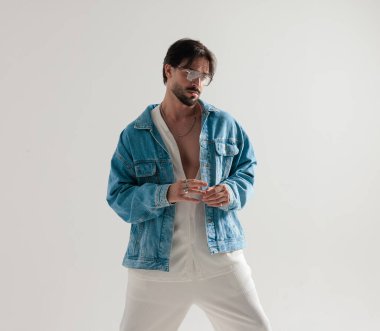 Kot ceketli yakışıklı adam parmaklarını bir arada tutuyor ve açık gri arka planda aşağıya bakıyor.