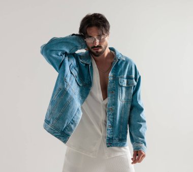 Kot ceketli sıradan bir adam boynuna dokunuyor ve açık gri arka planda dalgın dalgın bakıyor.