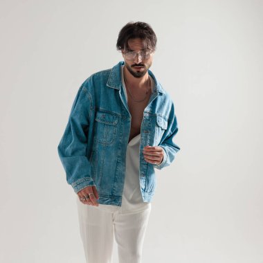 Moda adamı açık gri arka planda kot ceket ayarlarken ileriye bakıyor.