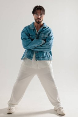 Kot ceketli ve beyaz pantolonlu ciddi ciddi sıradan bir adam kollarını kavuşturmuş açık gri arka planda bekliyor.