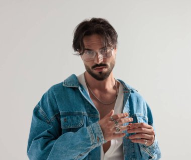 Kot ceketli moda adamı, elinde yüzüklerle açık gri arka planda dikilirken ileri bakıyor.