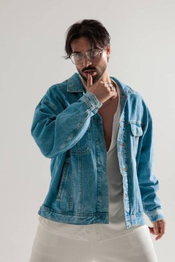 Kot ceketli dalgın bir adam parmaklarıyla dudağa dokunuyor ve açık gri arka planda ileriye bakıyor.