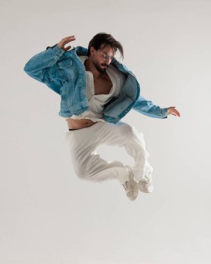 Kot ceketli havalı moda adamı havada zıplıyor ve açık gri arka planda dinamik bir şekilde aşağıya bakıyor.