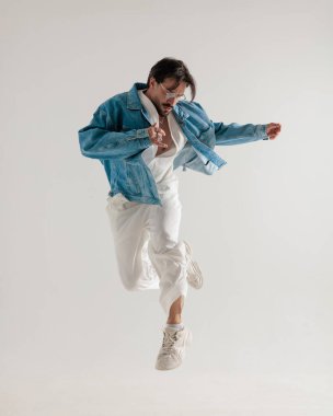 Kot ceketli sıradan şık bir adam açık gri arka planda havada zıplıyor ve poz veriyor.