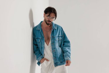 Kot ceketli sıradan bir moda adamı, açık gri arka planda el ele bekleyen beyaz bir duvara yaslanıyor.