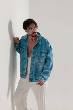 Kot ceketli zarif düşünceli adam beyaz bir duvara yaslanıyor ve açık gri arka planda diğer tarafa bakıyor.