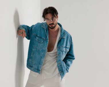 Kot ceketli moda adamı, bir eli cebinde beyaz bir duvara yaslanmış açık gri arka planda ileri doğru bakıyor.