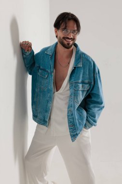 Kot ceketli, gülümseyen stil sahibi adam beyaz bir duvara yaslanıyor ve açık gri arka plana bakıyor.