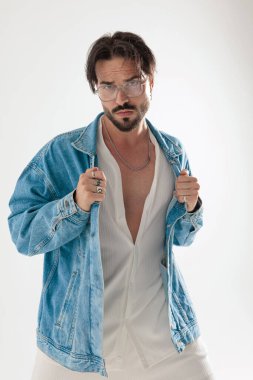 Beyaz arka plana bakarken kot ceket tutan ciddi sakallı bir adam.