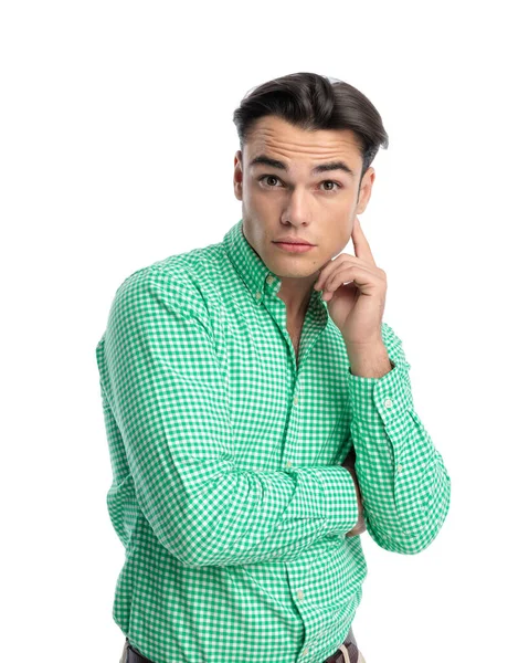 Yeşil kareli gömlek giyen, yüzüne dokunan ve beyaz arka planda ileriyi düşünen meraklı genç adam.
