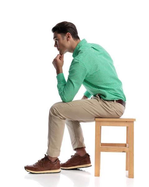 Tahta bir taburede oturan ve elleri çenesinde beyaz arka planda düşünen düşünceli genç adam.