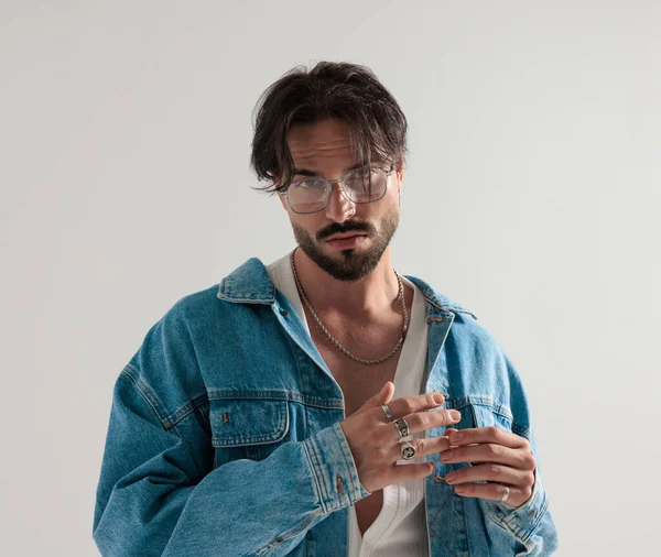 Kot ceketli moda adamı, elinde yüzüklerle açık gri arka planda dikilirken ileri bakıyor.