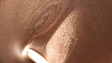 Koyu altın rengi boya, ahşap sopa sayesinde kıvrılıyor ve yayılıyor. Kum dokusunu, altın parçacıklarını ve gölge değişimlerini vurgulayan bir oyuk oluşturuyor..