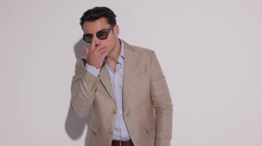 Havalı bir adam güneş gözlüğü takıyor, bej ceketi ilikliyor ve beyaz stüdyo arka planındaki kameraya bakarken el ele tutuşuyor..