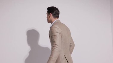 Bej takım elbiseli şık bir adam geri dönüyor, uzaklaşıyor, kameraya dönüyor, el ele tutuşuyor ve beyaz stüdyo arka planına yaslanırken ceketini ayarlıyor..