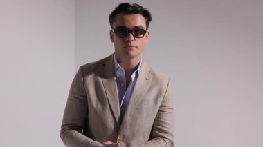 Bej takım elbiseli ve güneş gözlüklü ciddi bir adam elleri bağlı, onları ceplerine yerleştiriyor, beyaz arka planda dönüyor ve uzaklaşıyor..