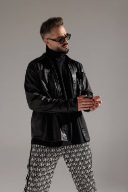 Siyah deri ceketli ve geometrik desenli pantolonlu şık genç adam gri stüdyonun arka grubuna bakarken el çırpıyor.