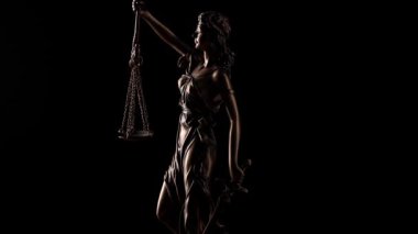 Roma tanrıçası Justitia 'nın gözleri bağlı figürü yavaşça yüzünü öne doğru çeviriyor. Kılıcı kavrıyor ve siyah bir arkaplana karşı yargı otoritesinin göstergesi olarak tartıyı tartıyor..