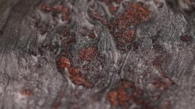 Koyu titanyum ve bakır boyanın detaylı makro görüntüsü yavaş hareket ederek, metalik yüzey boyunca organik, sıvı dalga desenleri yaratan kumlu bir dokuyu ve titreşen parçacıkları vurguluyor.