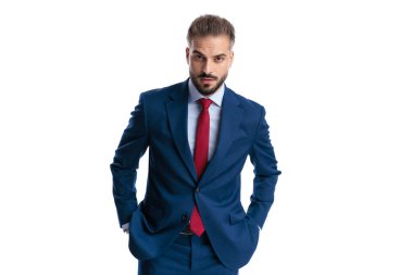 Serious man wearing blue suit and red tie, standing with hands in pockets and looking forward isolated on white background
