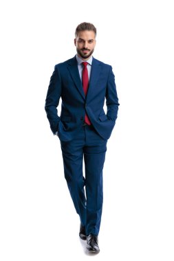 Confident man wearing navy suit walking forward with hands in pockets and looking forward on white background