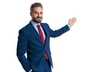 Confident man wearing blue suit presenting empty space to side with open palm while looking forward on white background