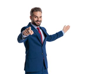 Happy businessman in a blue suit pointing a finger at the camera and presenting copy space with an open hand isolated on white background