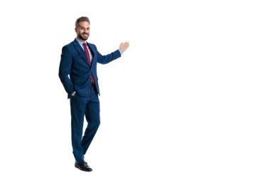 Inviting businessman wearing blue suit standing with hand in pocket and presenting blank space to side while looking forward on white background