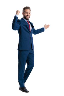 Excited businessman in a blue suit holding a fist up in victory and presenting copy space with an open hand isolated on white background