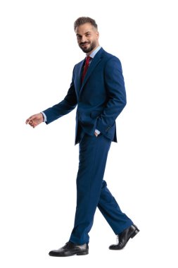Side view of elegant man in blue suit walking with hand in pocket and looking forward isolated on white background