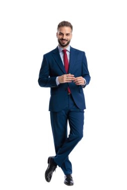 Confident man in suit rubbing hands and smiling while standing with crossed legs on white background