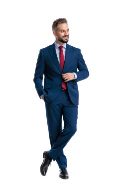 Elegant businessman wearing blue suit and red tie adjusting jacket and standing full body with crossed legs while looking side on white background