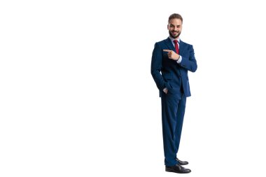 Mutlu iş adamı boş bir yeri işaret ediyor. Cebinde duruyor ve beyaz arka planda izole bir şekilde ileriye bakıyor.
