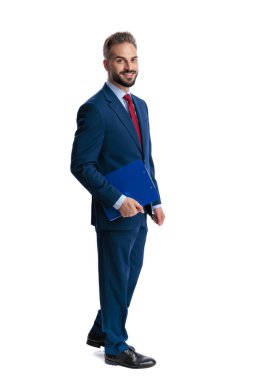 Side view of happy man in blue suit holding clipboard and smiling while walking isolated on white background