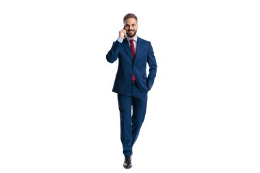 Happy businessman wearing a blue suit walking forward and talking on a smartphone with hand in pocket on white background